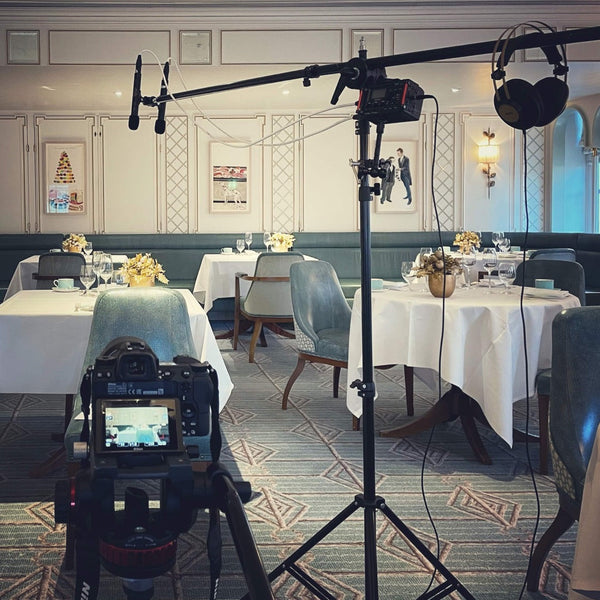 Picture of a tea room with light teal chairs at tables set with white tablecloths, and in the foreground is a camera facing the scene and to the right is a microphone stand with headphones at the near end of the boom pole and two black Sontronics STC-1S microphones at the far end of the boom pole