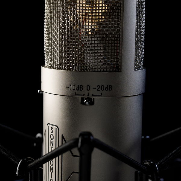 Close-up image on black background of the cylindrical body of a silver Sontronics STC-3X microphone with the silver pad switch in the centre and the text -10dB, zero and -20dB above it. Above this is the grille mesh through which the gold circular capsule is visible