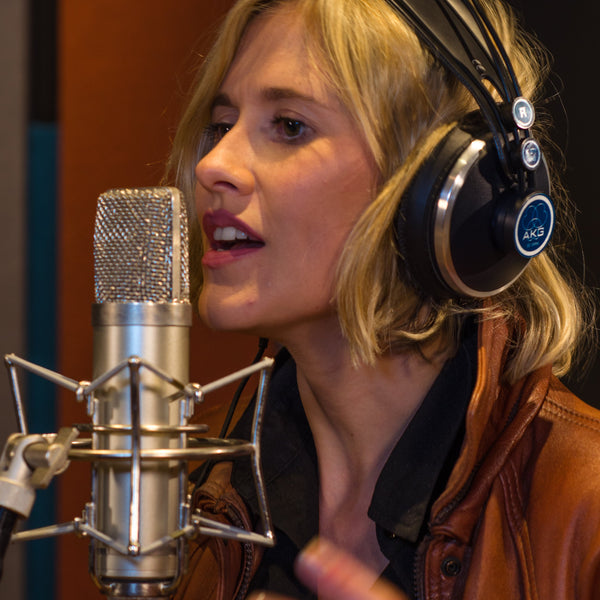 Close-up image in a recording studio of a woman with dark blonde hair wearing a pair of headphones with a black shirt and brown leather jacket singing into a Sontronics Aria microphone