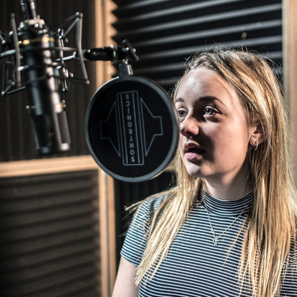 Image of a recording studio with acoustic panels in the background. On the right is a girl with long hair wearing a striped t-shirt, facing to the left and singing towards a black Sontronics STC-3X microphone which is suspended upside down in its spider shockmount and with the circular popshield between the mic and the girl's mouth