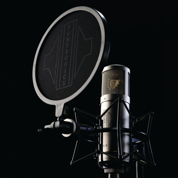 Image on black background with moody lighting of a large cylindrical Sontronics silver STC-2 microphone sitting in its spider shockmount facing slightly upwards to the left with the black circular nylon-mesh popshield in front of it showing the large white Sontronics logo 