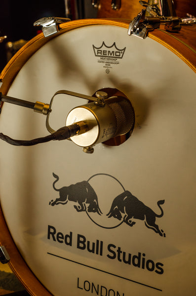 An image of the circular front head of a kick drum in its wooden frame and a Remo logo at the top. In to the circular soundhole is pointing the end of a Sontronics DM-1B microphone, suspended in its yoke mount from a microphone stand. Below this, the Red Bull Studios London logo and name is visible
