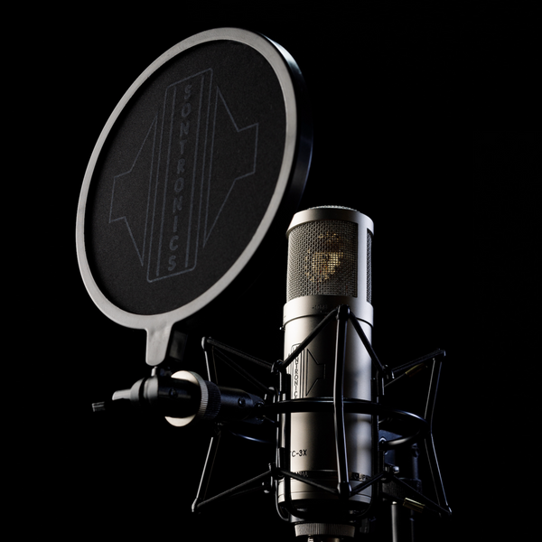 Image on black background with moody lighting of a large cylindrical Sontronics silver STC-3X microphone sitting in its spider shockmount facing to the left tilted upwards and with the black circular nylon-mesh popshield in front of it showing the large white Sontronics logo 