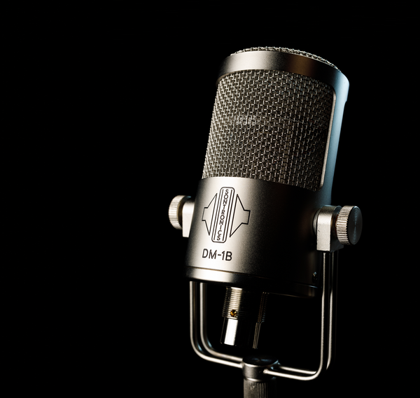 An image on black background with moody lighting showing the silver body of a sontronics DM-1B microphone in its U-shaped yoke mount pointing to the left and tilted slightly upwards and away from us