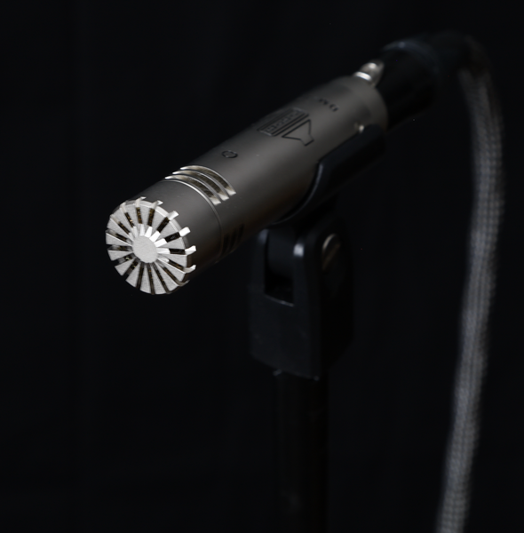 An image of a silver-coloured Sontronics DM-1T microphone mounted in its clip with a corded cable attached facing towards us from out of a black background
