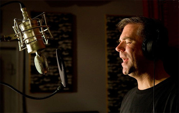 An image of a recording studio with acoustic panels and red curtain on the back wall. In the foreground on the right is a man with short hair wearing a black t-shirt and headphones facing to the left and singing towards a Sontronics Mercury microphone, which is hanging upside down from a mic stand, facing towards the man and with a Sontronics ST-POP popshield in front of it