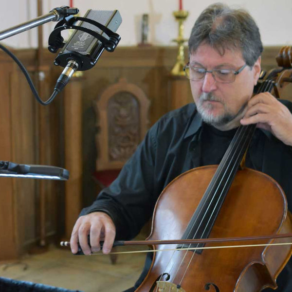 image of a a man with glasses and a beard in a black shirt playing a cello in a church setting. To the top left of the picture is a Sontronics Sigma 2 microphone suspended in its shockmount from a microphone stand, pointing down towards the cello