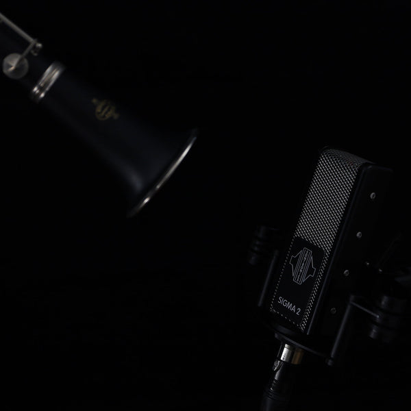 An image on black background with moody light of the bell of a clarinet coming in from the top left corner and in the bottom right corner, pointing up towards the clarinet, is a Sigma 2 microphone in its shockmount