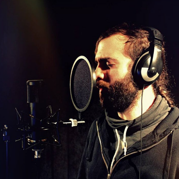 Image on a black background of a man with a beard and long hair and closed eyes, wearing a hoodie, facing to the left and singing into a circular black popshield that sits 10 centimetres in front of a black Sontronics STC-20 microphone in its spider-style shockmount mounted on a microphone stand